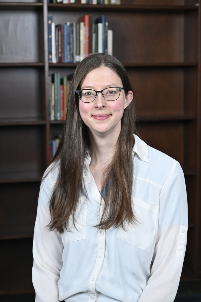 Portrait of Jane LaBarbara taken in Wise Library at West Virginia University. Jane is seated wearing a white button down a partially filled bookcase is in the background.